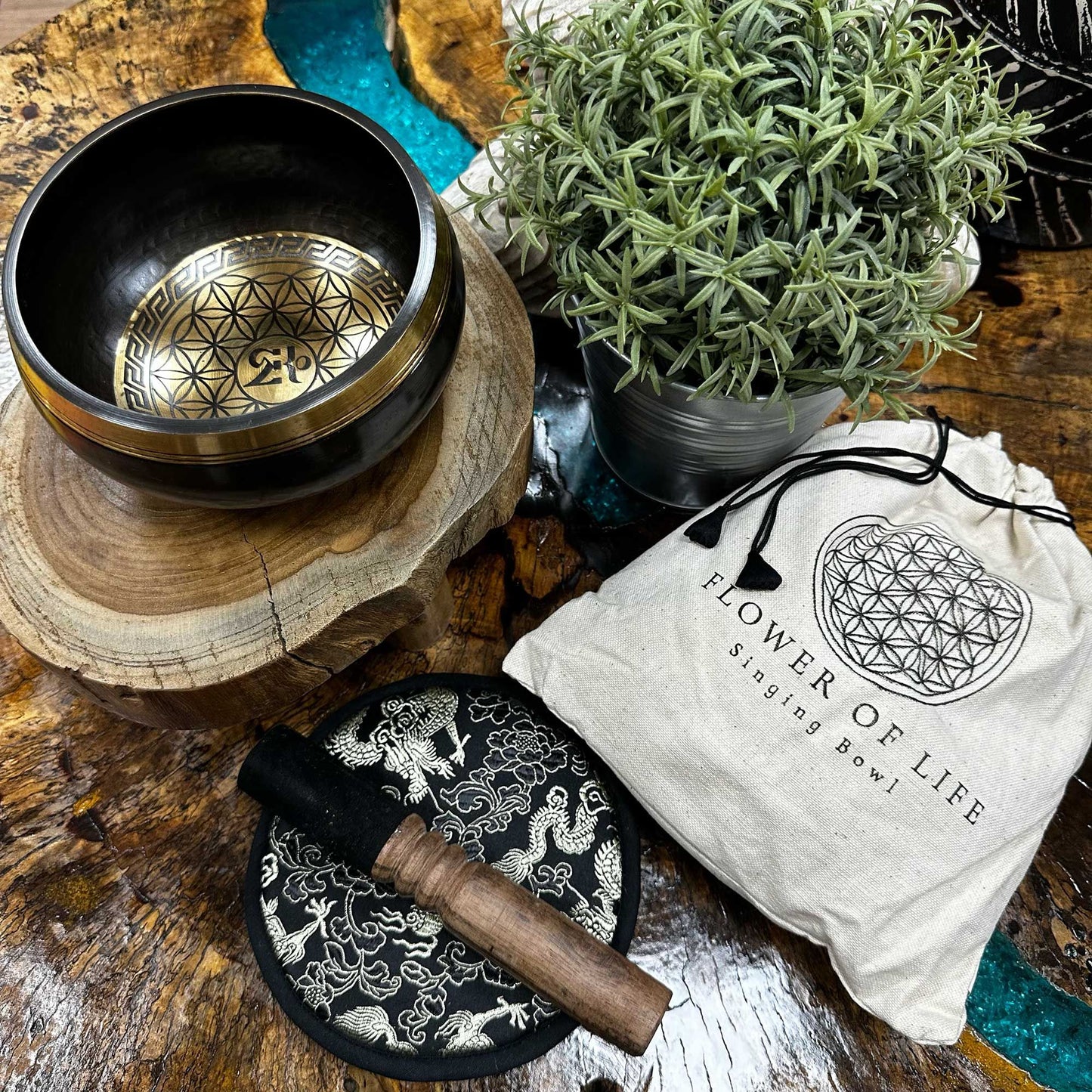 Singing bowl sits on a wooden slab among a mallet on a decorative pad and a potted plant beside a cloth bag labeled "FLOWER OF LIFE Singing Bowl" on a rustic table.
