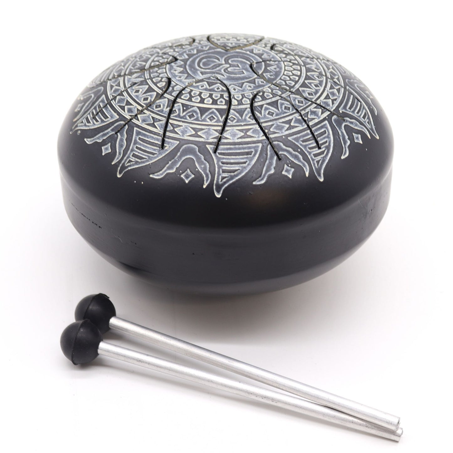 Black steel tongue drum with white etched mandala and scored tongues rests on a white surface while two metal mallets with rubber tips lie beside it.