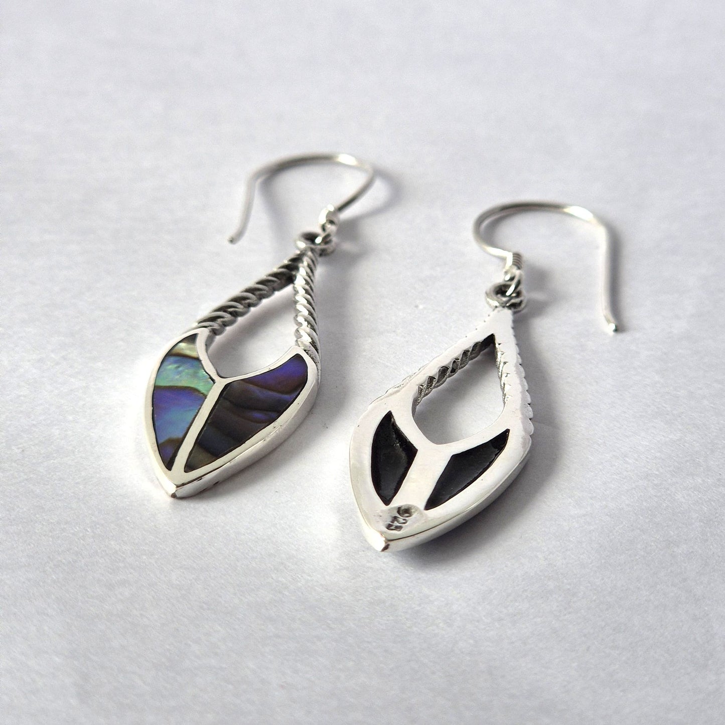 Silver teardrop earrings with twisted stems, one showing iridescent abalone inlay and the other black inlay stamped 925, lying on a white paper surface in close-up.