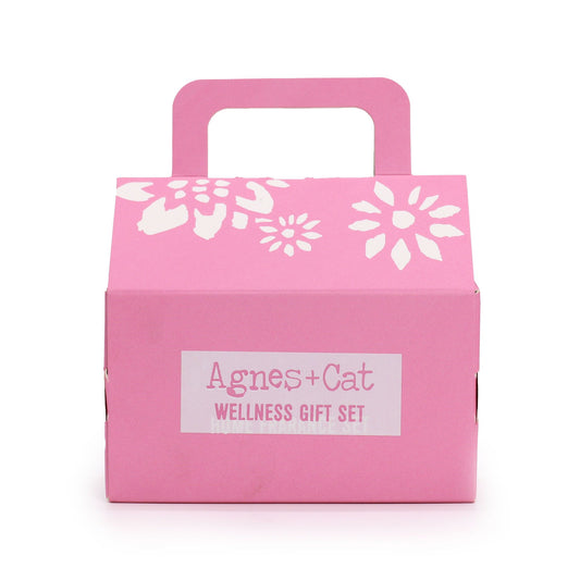 Pink cardboard gift box sits closed with a carry handle, decorated with white floral prints and labeled Agnes+Cat WELLNESS GIFT SET against a white background.