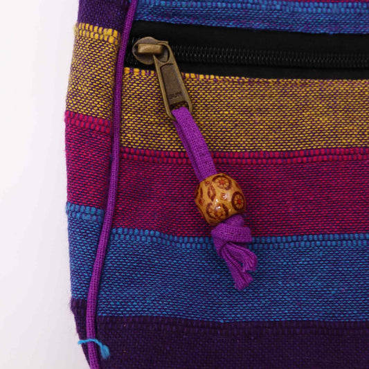 Striped woven pouch showing a brass zipper pull attached to a purple cord threaded through a carved wooden bead against horizontal bands of yellow, magenta, turquoise and navy fabric