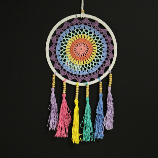Colorful crocheted circular dreamcatcher hangs from a white cord, showing concentric pastel rings and seven tasseled cords with wooden beads against a plain black wall.