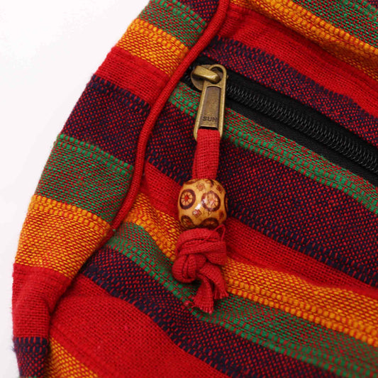 A brass zipper pull stamped "SUN" hangs from a red fabric cord with a carved wooden bead, attached to a black zipper on a red green and yellow striped bag.