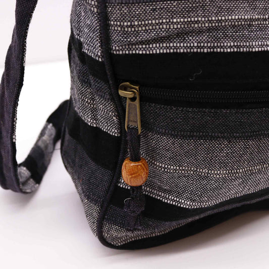 Small gray and black striped woven shoulder bag displays a front zippered pocket with a brass pull labeled SUN and a black cord ending in a wooden bead, resting on a white surface.