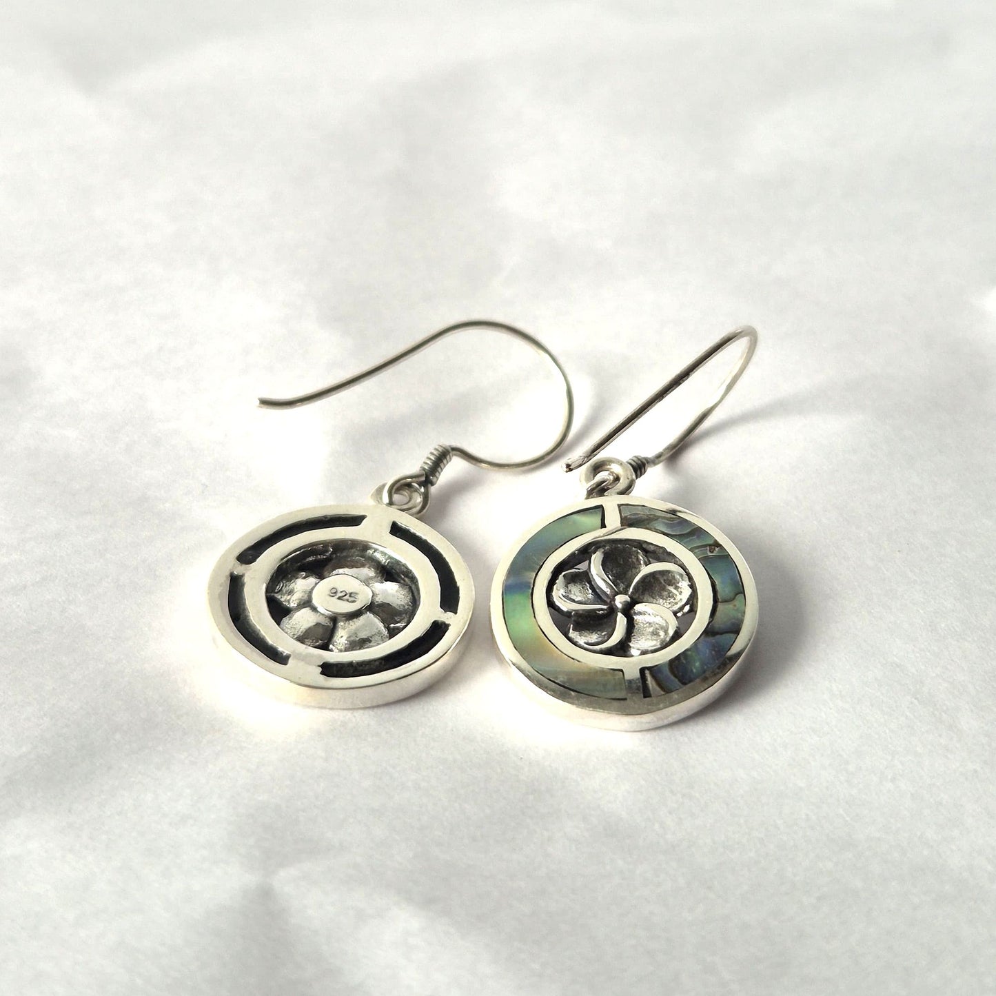 Silver circular drop earrings rest on crumpled white paper; each has a floral motif and abalone inlay, the left stamped "925" center, curved ear wires casting soft shadows.