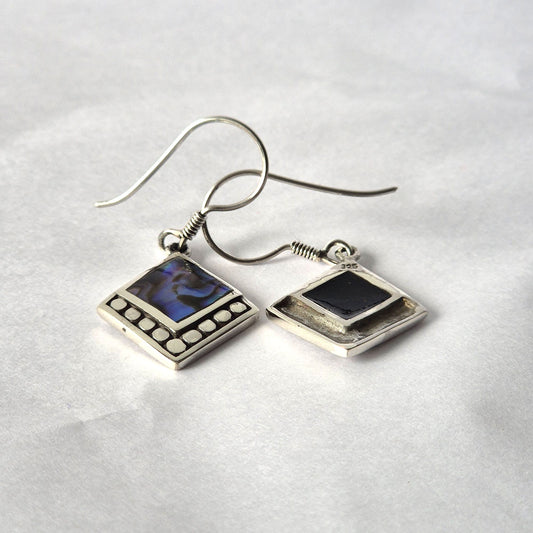 Pair of silver dangle earrings featuring iridescent abalone squares and dotted borders, lying with intertwined hooks on white paper backdrop, one earring back stamped 925.