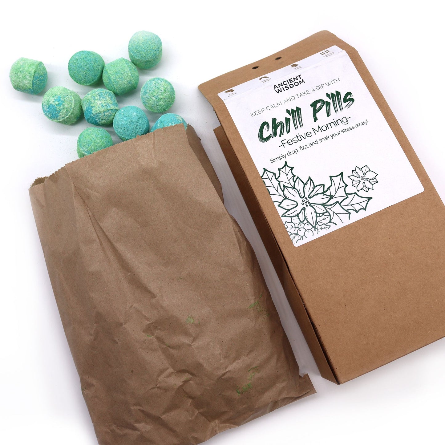 A cluster of small green-blue spherical bath bombs spilling from a torn brown paper bag next to a brown cardboard box on a white surface; label text: ANCIENT WISDOM KEEP CALM AND TAKE A DIP WITH Chill Pills -Festive Morning- Simply drop, fizz and soak your stress away!