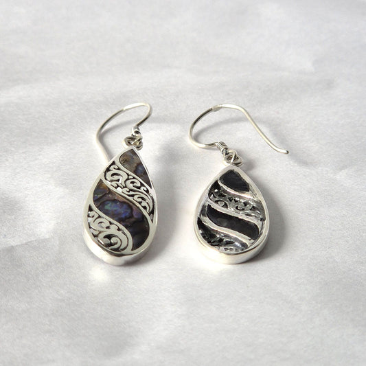Teardrop silver earrings rest side by side; one features iridescent abalone with ornate scroll filigree, the other darker textured silver; both with fishhook wires on crumpled white paper.