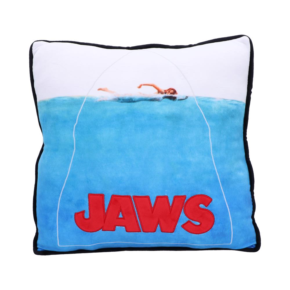 A square pillow, trimmed in black, features the poster art from the movie "JAWS" in which a swimmer is shown above the surface and the word “JAWS” in red below, with the silhouette of a shark connecting them.