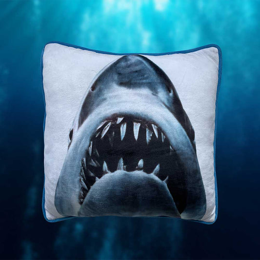 A blue-trimmed white pillow displays the upward-facing head of a shark with its mouth open, revealing rows of teeth, against a blurry, blue background.