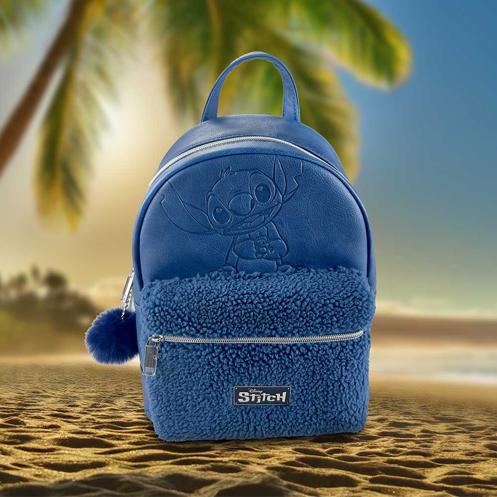 A blue Stitch backpack sits on a sandy beach. The backpack features an embossed Stitch image and a fuzzy bottom section. Disney Stitch is printed on a small metal plate.