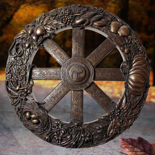 A bronze wheel decorated with fruits, leaves, and symbols is inscribed with "Beltane," "Yule," and other seasonal names. It's displayed in an outdoor autumn setting, surrounded by fallen leaves.