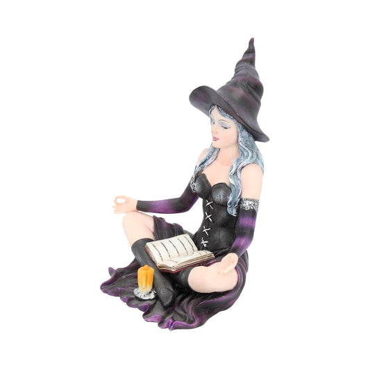 A figurine of a witch with a pointed hat sits cross-legged, meditating with closed eyes, a book on her lap, and an unlit yellow candle beside her on a mat.