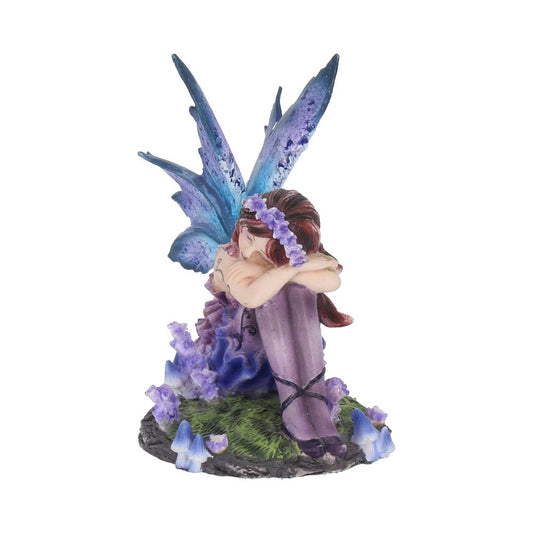 A delicate fairy figurine, with blue wings and purple attire, sits with knees drawn up, surrounded by grass and blue flowers, creating an enchanting, mystical scene.