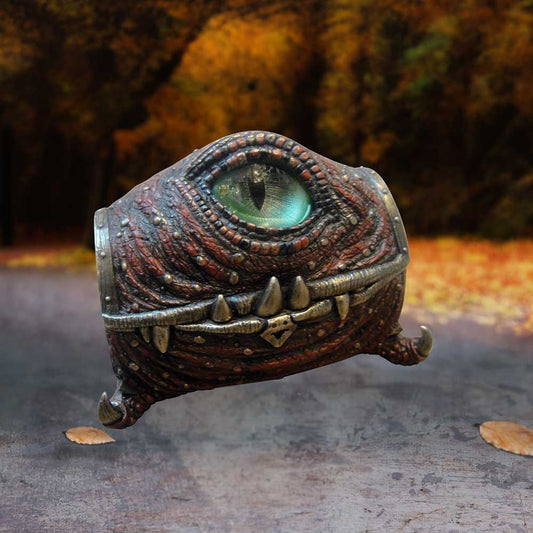 A textured creature with a large, single eye and sharp teeth peers forward. It appears on a path surrounded by blurred autumn foliage, creating an eerie, mystical atmosphere.