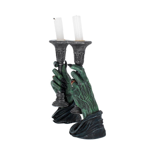 Green gnarled hands clutch two ornate silver candlesticks with unlit white candles, hands emerging from dark tattered sleeves against a plain white background.