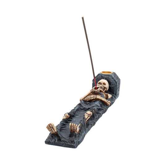 A skeleton incense holder, resting on a dark surface, burns an incense stick.