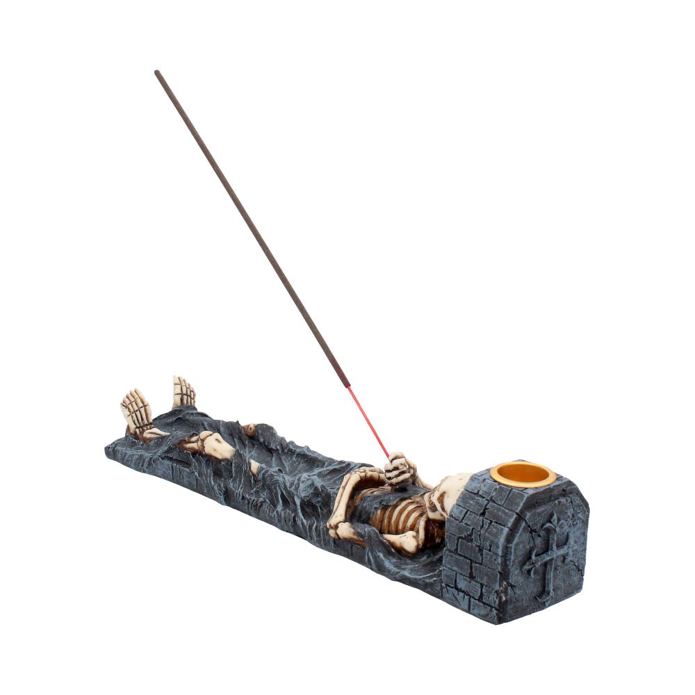 A skeleton figurine lies on a dark gray incense burner; a long incense stick burns, resting in the holder. The scene is isolated on a white background.