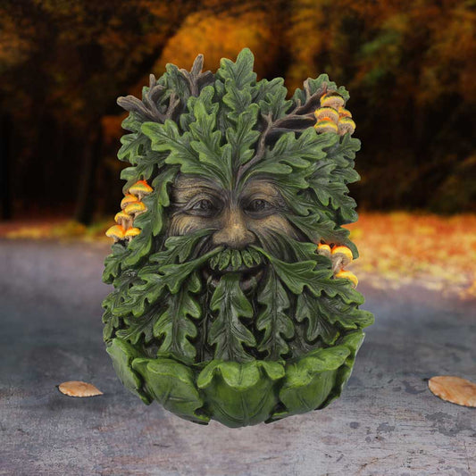 Green, leafy face sculpture with oak leaves and orange mushrooms, smiling. The background features an out-of-focus forest with autumn colors and scattered leaves on a gray path.