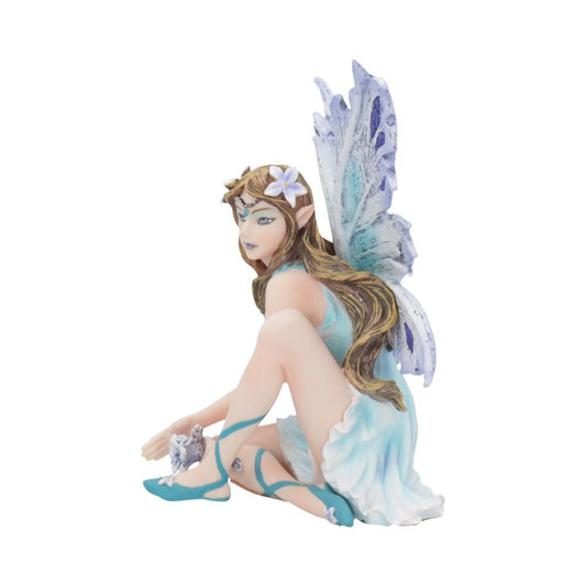 A fairy figurine with pastel wings and long hair kneels, wearing a blue dress, holding a small creature. The background is plain and white, providing a minimalist setting.