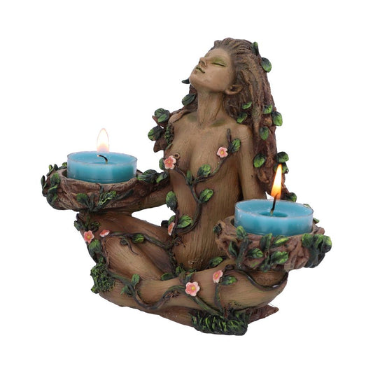 A wood-textured tree-woman statue sits cross-legged, vines and pink flowers wrapping her body as she holds two lit blue tealight candles in rustic bark bowls against a white background.