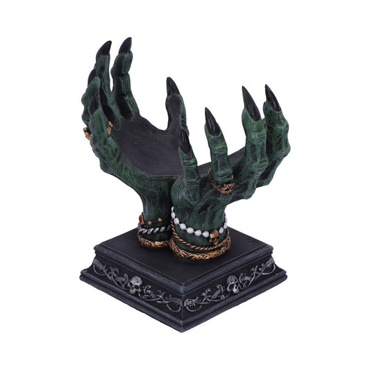 Two green, clawed witch hands are cupped upwards, supporting a dark platform. The base of the hands are adorned with jewelry and sit upon a square base decorated with skulls.
