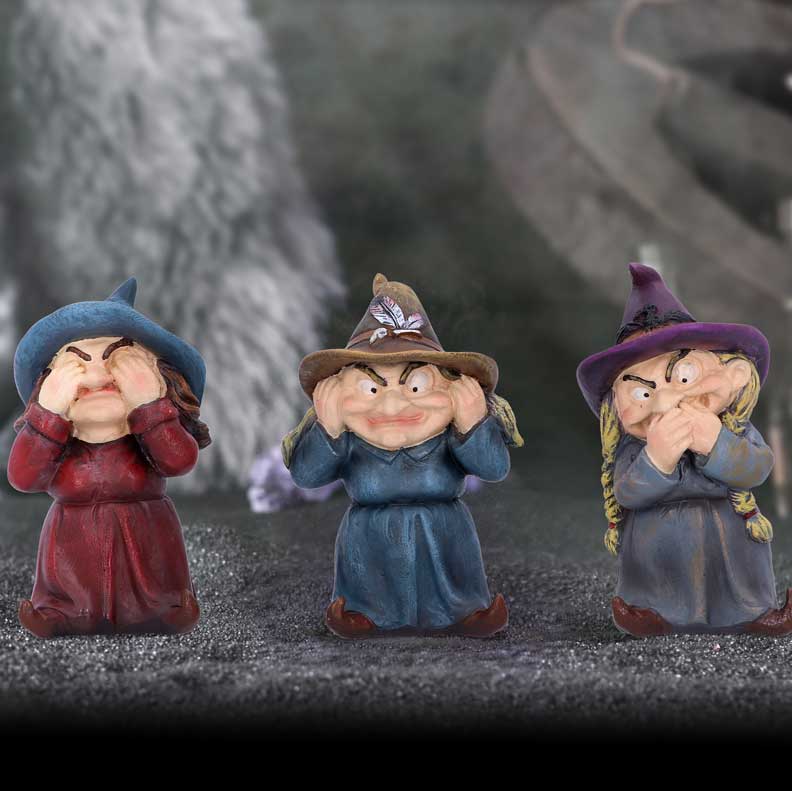 Three small figurines of witches, each in different poses resembling "see no evil, hear no evil, speak no evil," stand on a textured surface. The background is dark and slightly blurred.