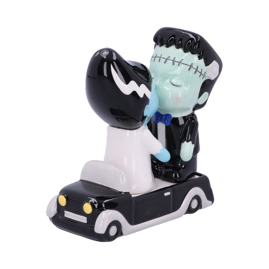 A Frankenstein and his bride figurine kiss atop a black car with yellow headlights, set against a plain white backdrop.