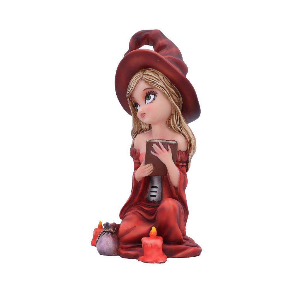 A figurine of a young witch in a red hat reads a book while kneeling. She is surrounded by lit candles and a small sack, on a plain white background.