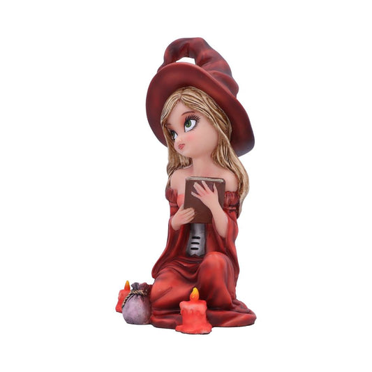 A figurine of a young witch in a red hat reads a book while kneeling. She is surrounded by lit candles and a small sack, on a plain white background.