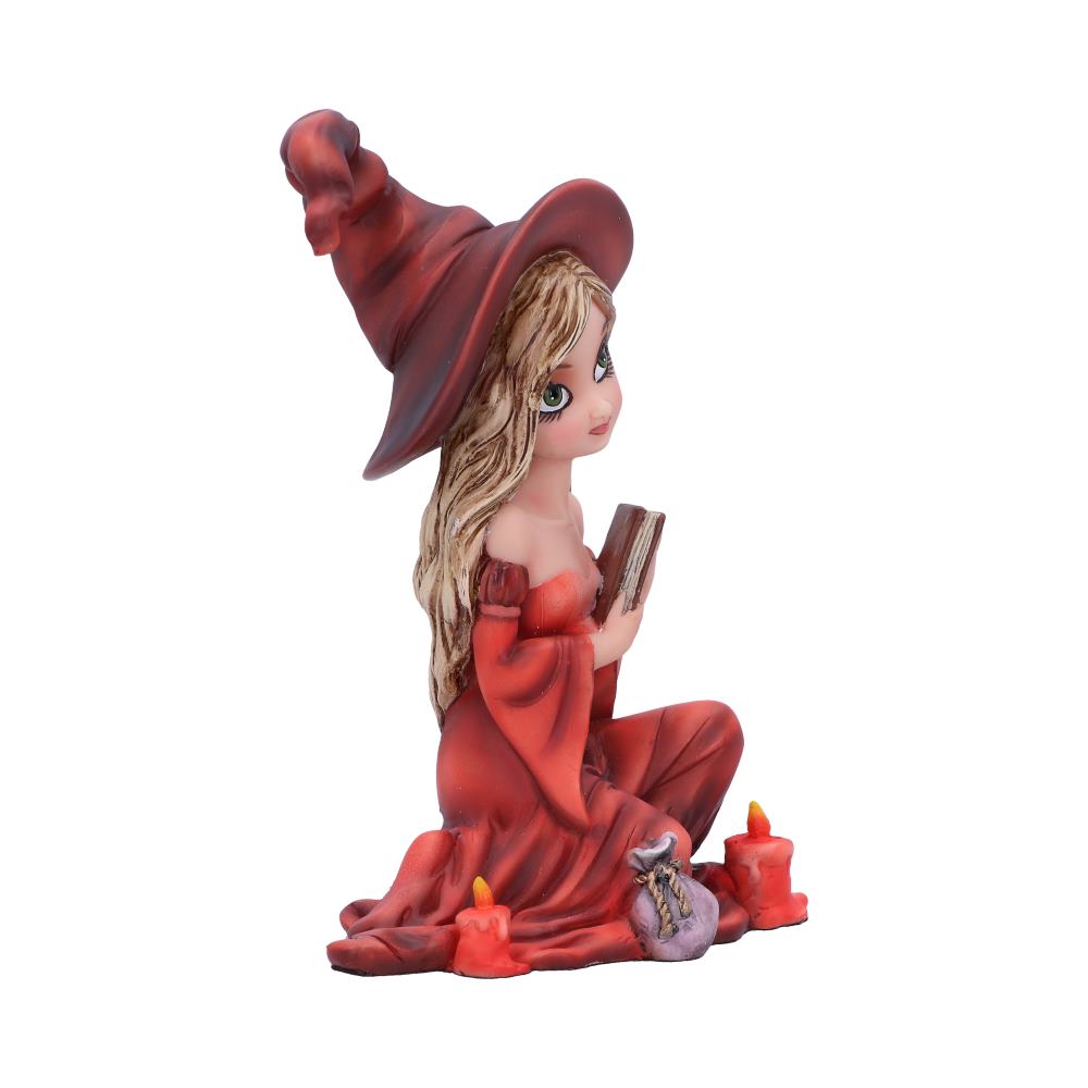 A figurine of a young witch wearing a tall, pointed hat and red dress sits while holding a book. She is surrounded by lit candles and a small bag, all against a plain background.