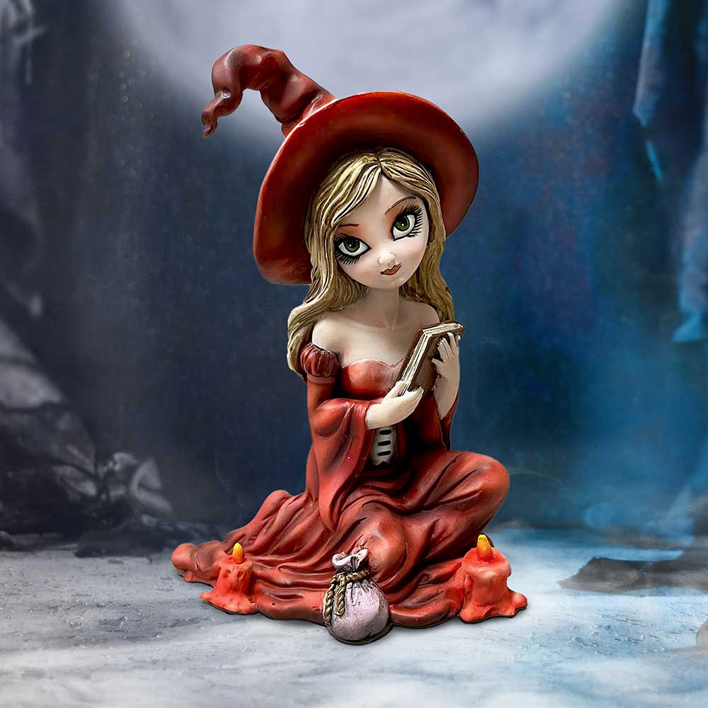 A clay figure of a young witch, wearing a red dress and hat, holds a book. She sits cross-legged on a stone floor, surrounded by candles and a small pouch.
