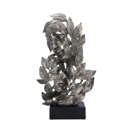 A metallic sculpture depicts an abstract human face intertwined with detailed leaves, creating a textured, leafy pattern mounted on a simple black base.