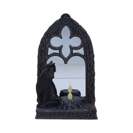 A gothic-style mirror framed in sculpted branches shows a black cat sitting beside a small, flickering faux candle. The mirror is shaped like a quatrefoil and topped with a pentagram.