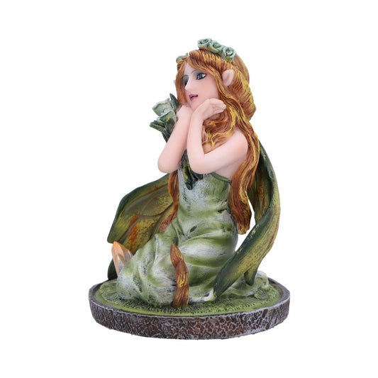 A figurine of a fairy sits; she is wearing a green dress and has long auburn hair, resting her chin on her hands. The fairy is situated on a circular base resembling a tree stump.