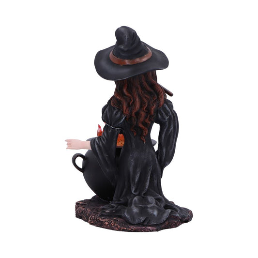 A witch figurine stirs a bubbling cauldron, wearing a wide-brimmed black hat and flowing black dress, long brown hair cascading down onto a textured base.