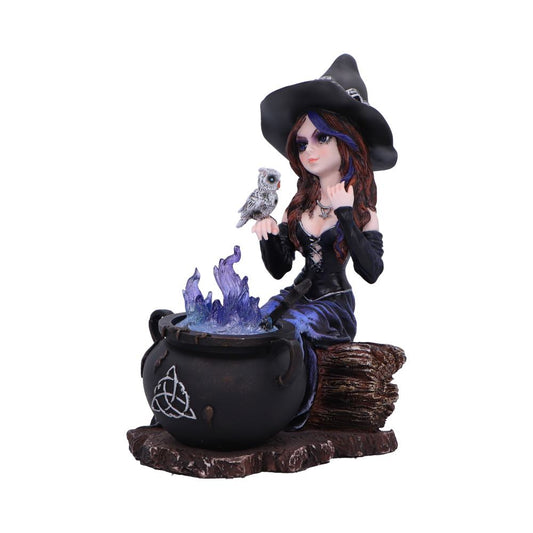Witch figurine wearing a black hat and purple dress sits on a log holding a small owl while stirring a black cauldron emitting purple blue flames on a wooden base.