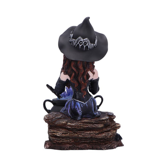 Witch figurine sits on a stacked log base stirring a glowing purple cauldron, wearing a black pointed hat and purple dress with long red hair against a plain white background.