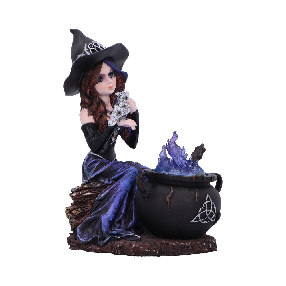 Witch figurine holding a small owl, seated on a log beside a cauldron emitting purple flames, wearing a black hat and purple gown, photographed against a plain white background.