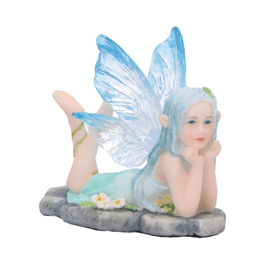Small fairy figurine reclining, chin in hands and legs kicked up, translucent blue wings and pastel dress resting on a stone slab adorned with tiny white flowers against white background
