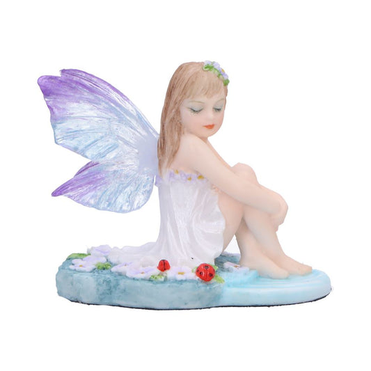 Fairy figurine sits with knees drawn to chest, iridescent purple-blue wings folded, wearing white dress and flower crown, perched on a blue floral base with tiny ladybugs.