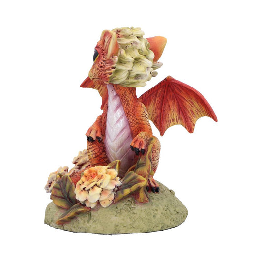 Orange-red baby dragon figurine sits on a textured green mound, wings partially spread, head tilted upward, pinkish scaled belly visible, surrounded by pale yellow-pink flowers and green leaves.