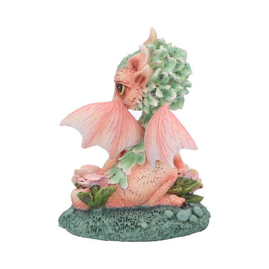Pastel baby dragon figurine sits curled, turning its head to the side, wings partially raised, perched on a textured green base with small leaves and flowers against a white background.