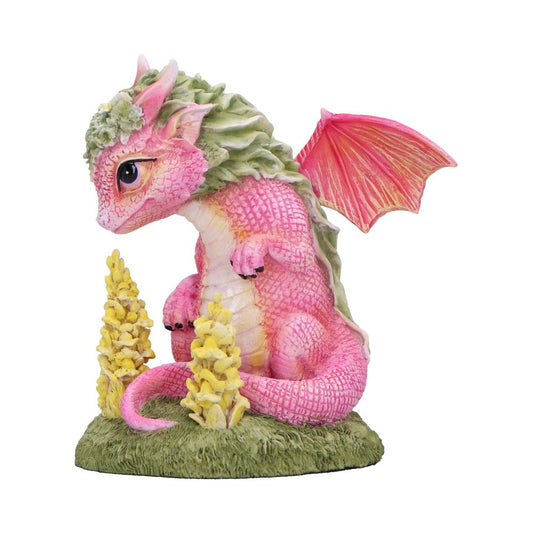 Pink baby dragon figurine sits curled on a grassy base, gazing downward beside two yellow flower spikes against a plain white studio background.