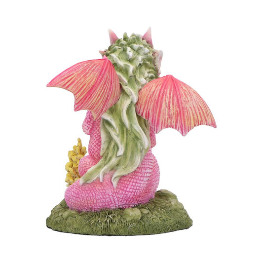 Pink dragon figurine sits facing away with outstretched pink wings, green mane flowing down its back, holding small yellow flowers while seated on a grassy base against a white background.