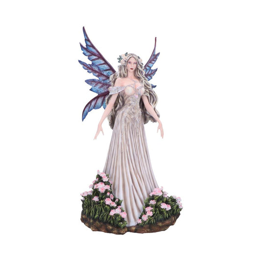 A porcelain fairy figurine stands with arms outstretched, wearing a flowing pale gown and a flower crown, iridescent blue-purple wings rising behind, rooted on a grassy base with pink flowers.