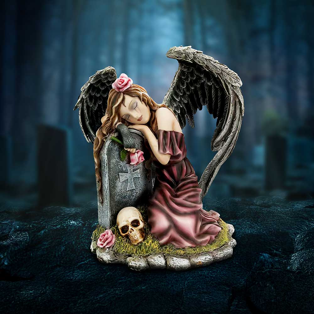 A winged female angel statue rests her head on a gravestone, surrounded by roses and a skull, wearing a pink dress with dark feathered wings in a misty forest cemetery.