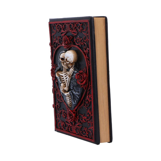 Hardcover book stands upright, its black and red embossed cover showing two embracing skeletons in a heart-shaped frame surrounded by roses and baroque scrollwork against a plain white background.