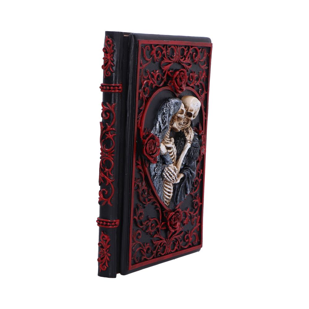 Gothic hardback book with ornate black and red embossed cover featuring a three-dimensional sculpture of two skeletons embracing and kissing inside a heart-shaped frame, photographed standing against a white background.
