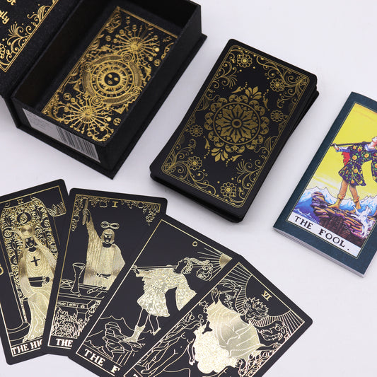 Tarot cards, featuring gold designs on black, are displayed with The Fool card, showing a figure on a cliff. The cards lie next to a box, suggesting a mystical theme.